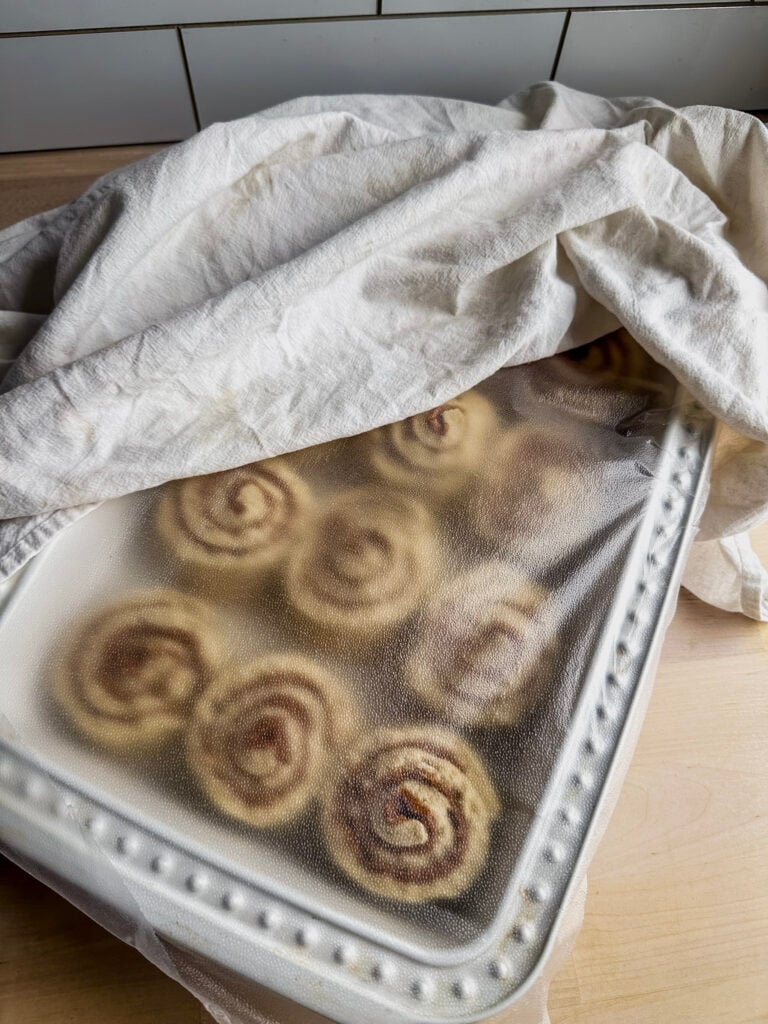 white 9 x 13 baking dish filled with gf cinnamon rolls ready to proof covered with plastic wrap and towel