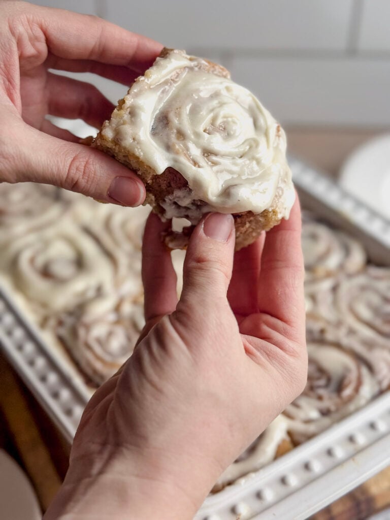 baked and frosted gluten free cinnamon roll being pulled apart by hands