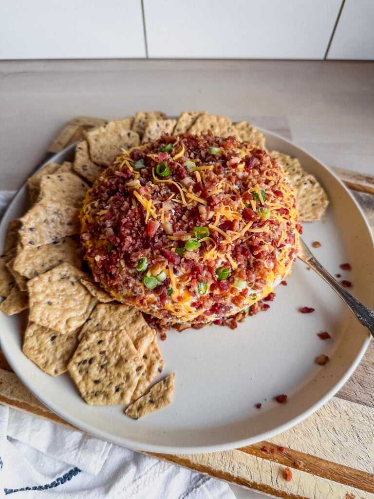 bacon covered cheese ball with green onions and shredded cheddar cheese on top of a serving plate with crackers on the side