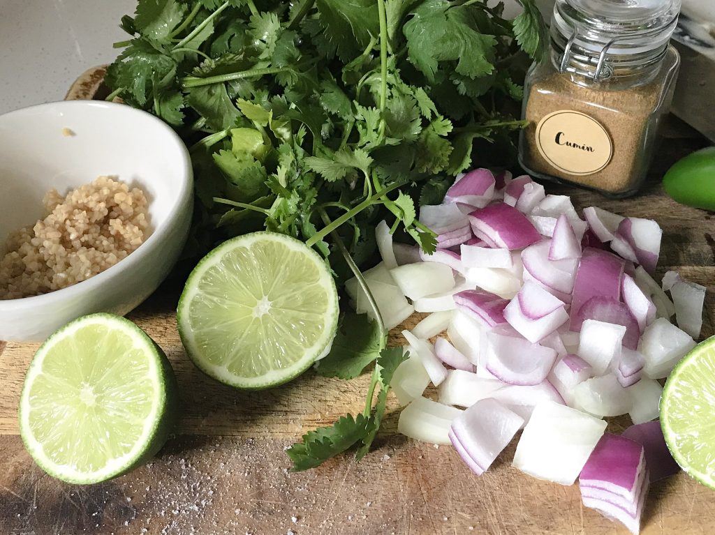 garlic, limes, cilantro, red onion and cumin sit atop a wooden cutting board