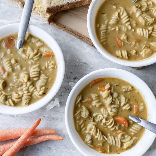 three bowls of gluten free chicken noodle creamy soup on table with carrots nearby