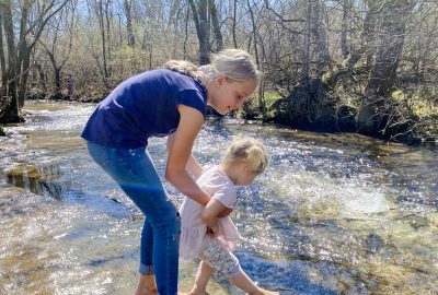 older girl helping younger girl play near a stream