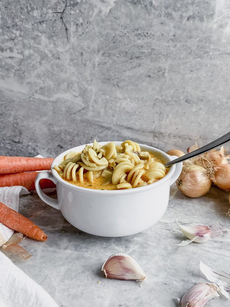 gluten-free creamy chicken noodle small bowl of soup with gray backdrop including carrots, onions and garlic