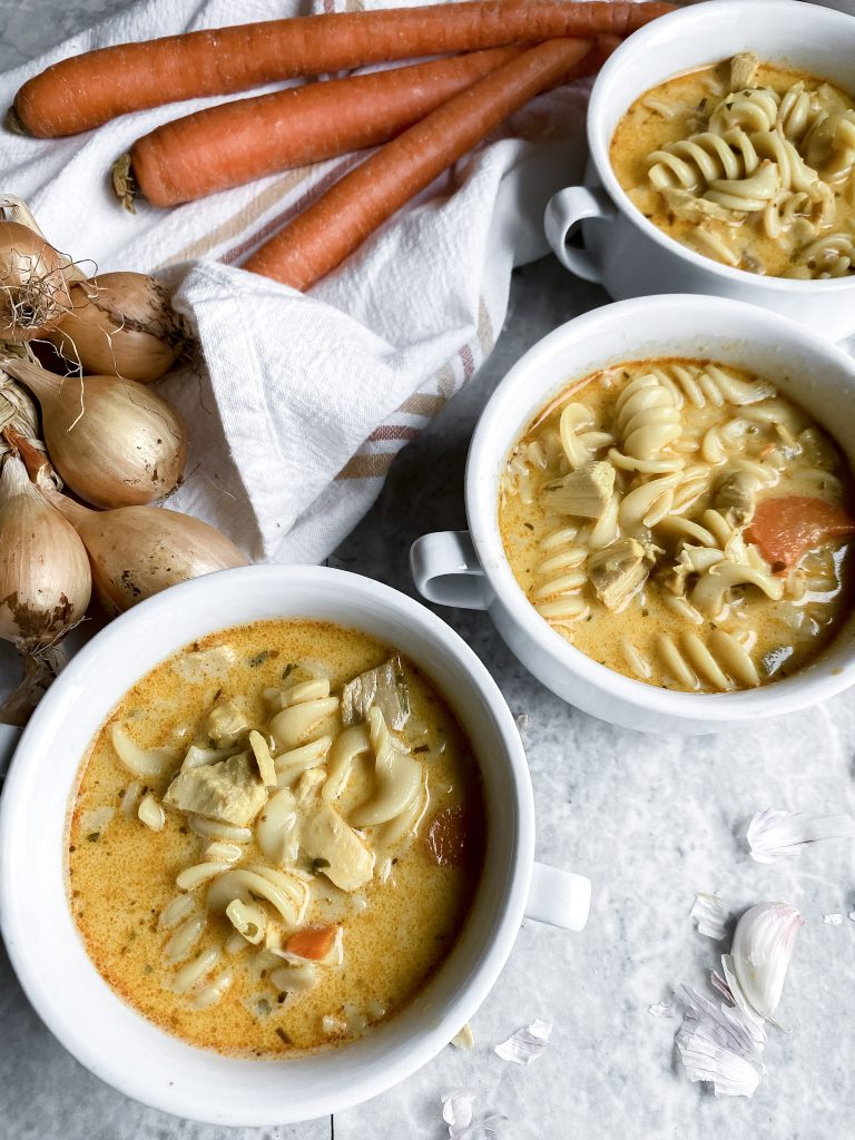 three bowls of creamy gluten free chicken noodle soup near each other as well as onions and carrots in background