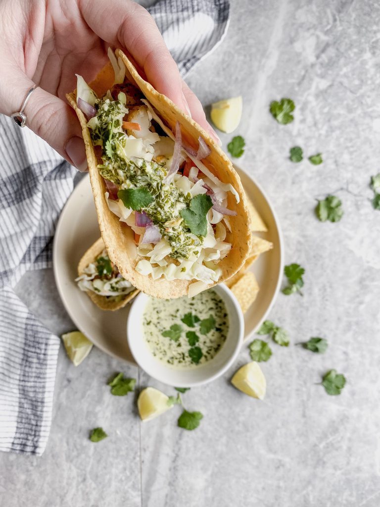 fish taco in corn tortilla with cilantro lime cream sauce being held above a gray surface