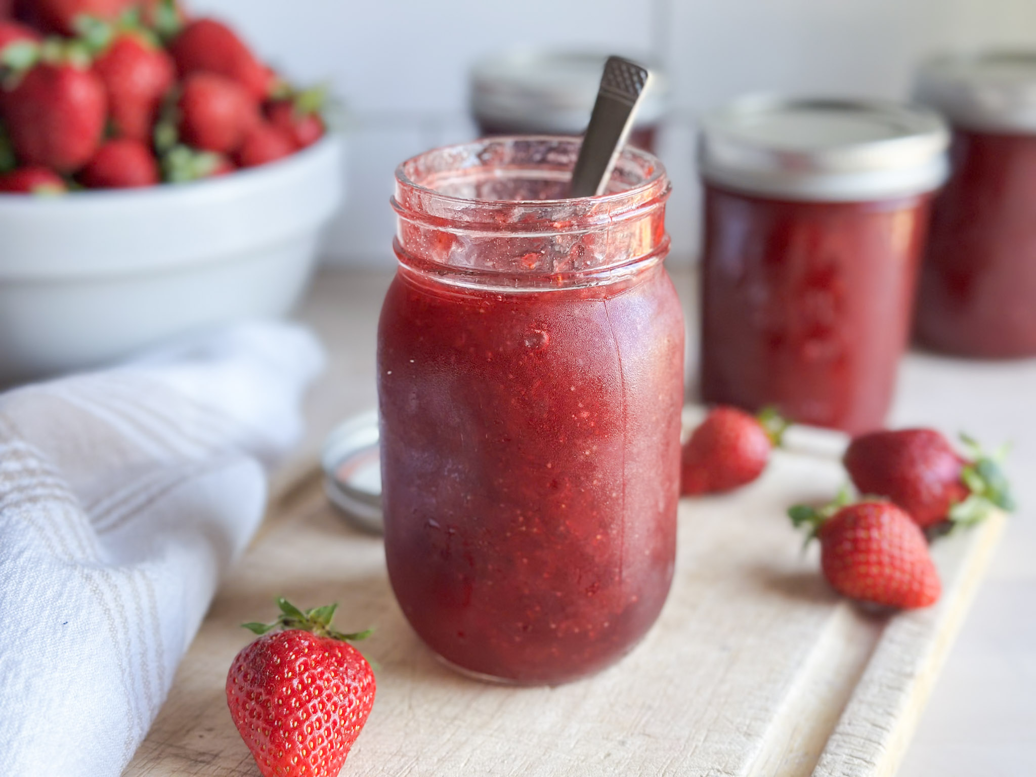 jar of homemade strawberry jam on top of a cutting board with strawberries in the background