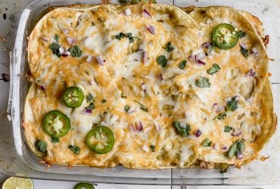 casserole dish holding a pan of gluten free green chicken enchiladas on a counter top with jalapeños, onions and cilantro sprinkled on top