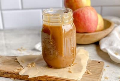 a jar of homemade caramel sauce on wooden cutting board with a bowl of red apples behind it.