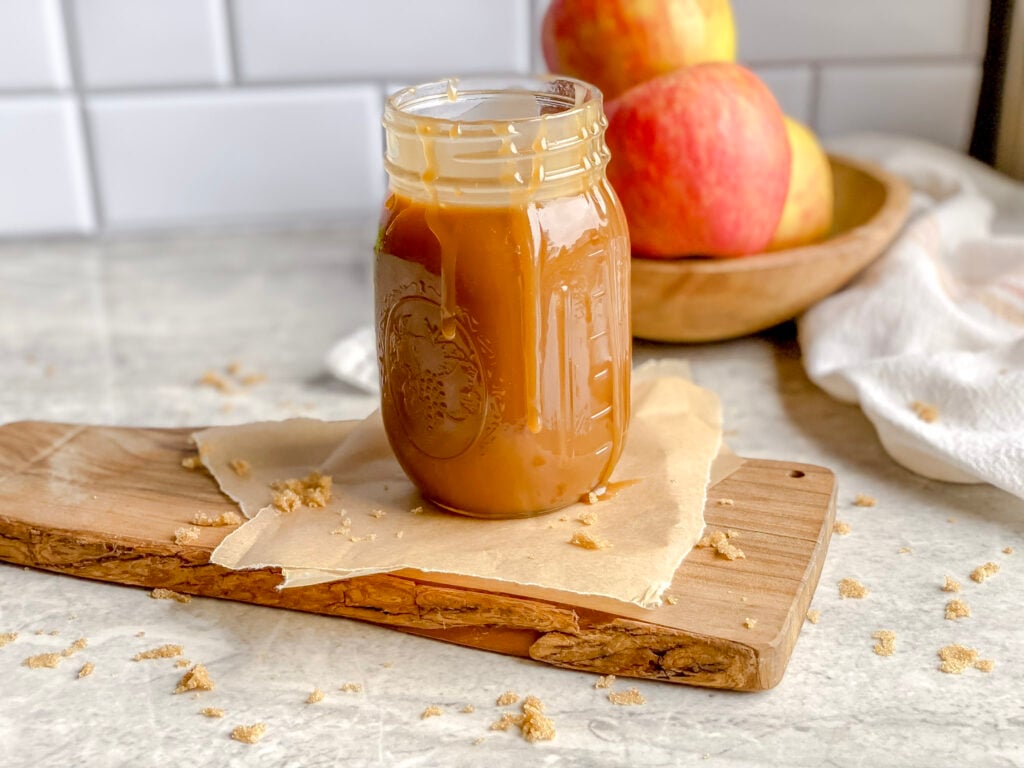 jar of deep brown caramel sauce on a wooden cutting board with red apples in the background