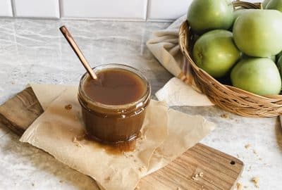 homemade vegan and dairy free caramel sauce in a jar sitting on parchment papers on top of a cutting board with green apples in a basket behind them