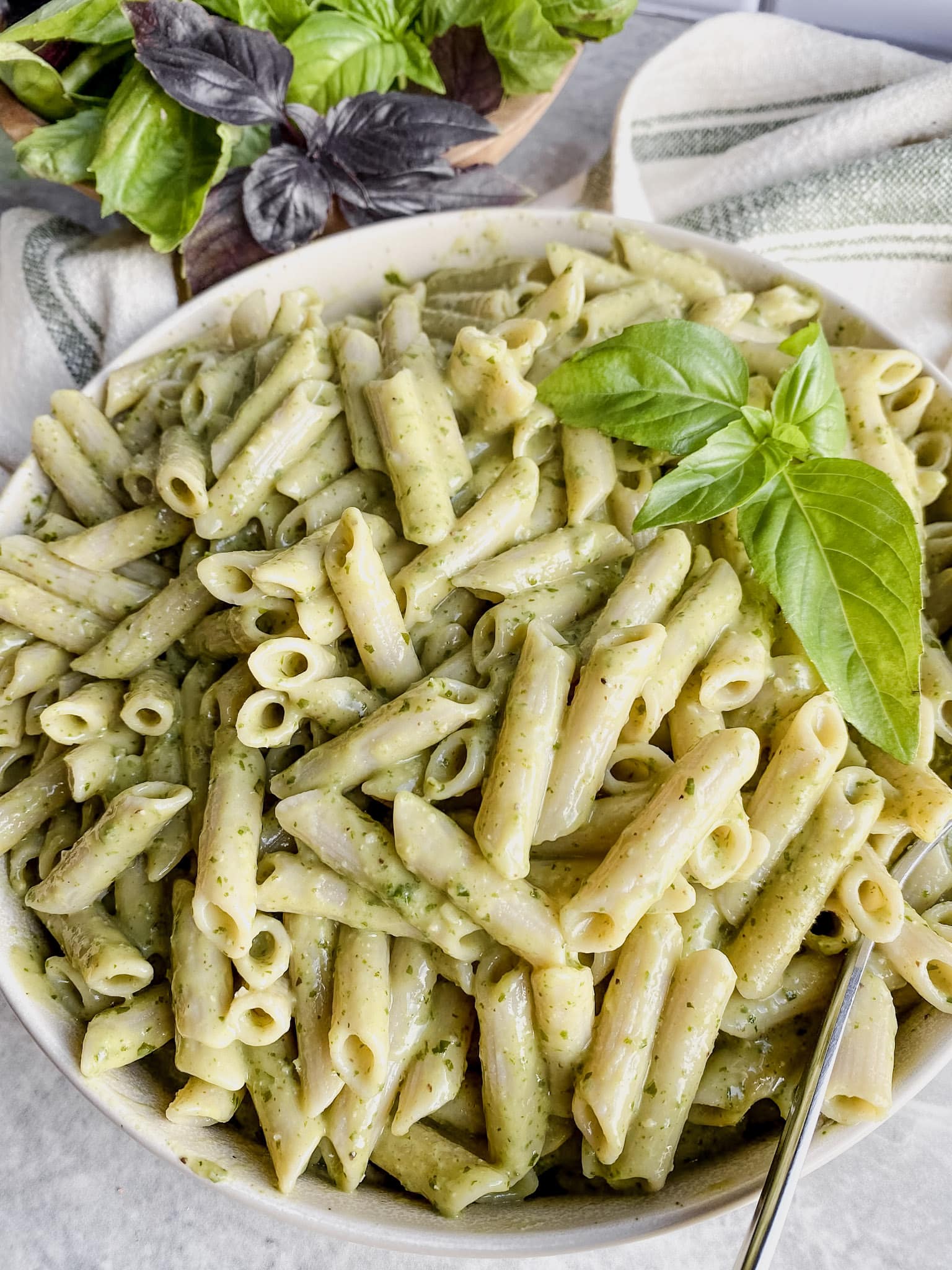 large bowl of gluten free penne pasta with dairy free pesto pasta sauce and fresh basil leaves on top