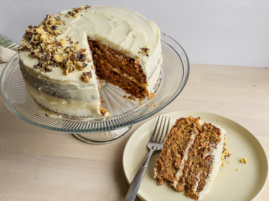 double round gluten free carrot cake with cream cheese frosting on a glass cake stand and a piece on the table