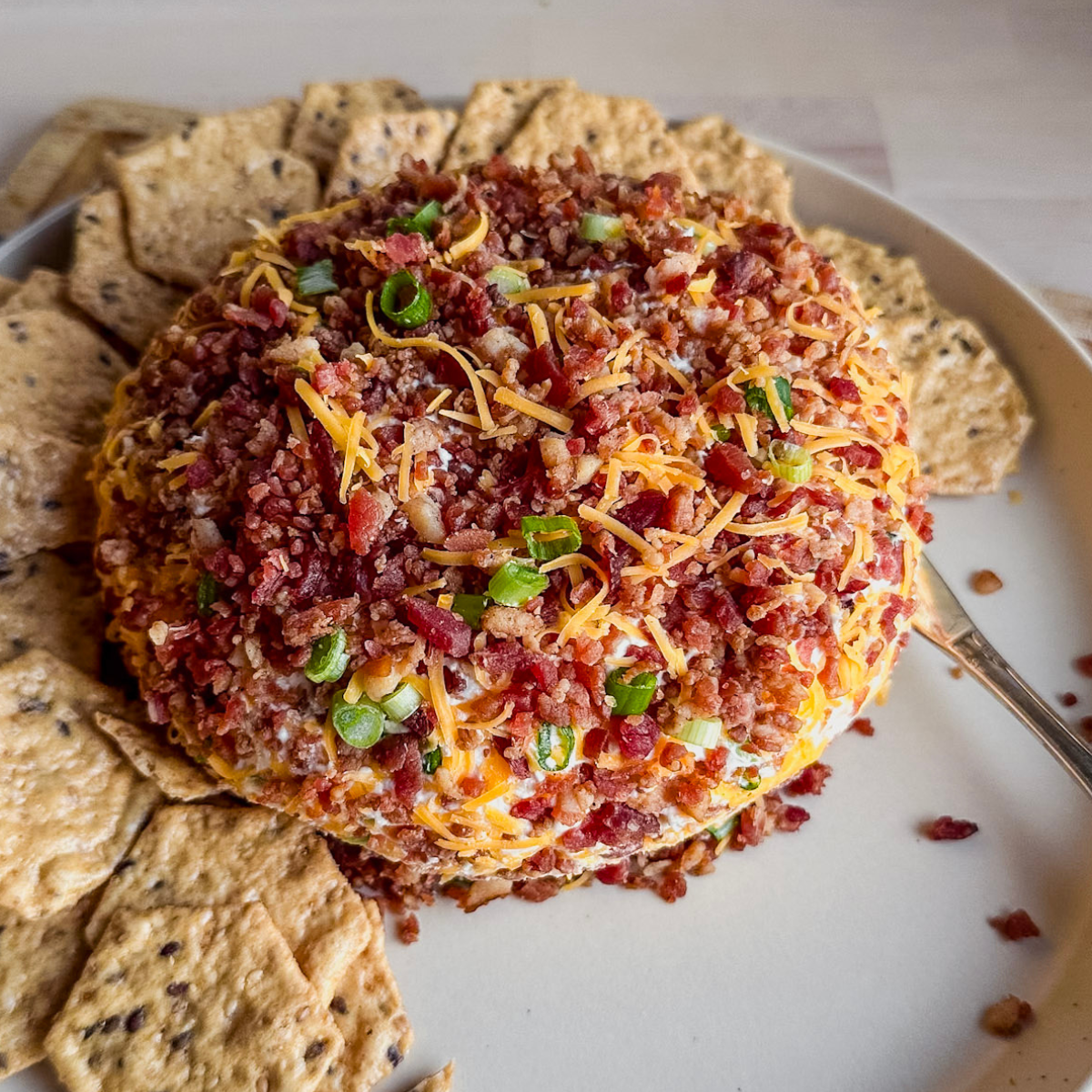 bacon bit covered cheese ball on a plate with gluten free crackers and a knife in the cheese ball