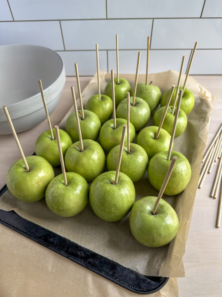 lined tray with parchment paper and green granny smith apples lined on top of it with wooden dowels sitcking out